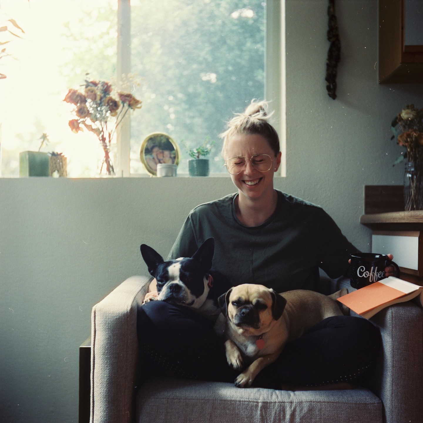 A woman sits in a chair with two dogs on her lap, holding a coffee mug and smiling.