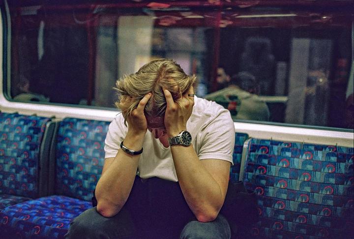 A man sits on a train with his head in his hands, looking distressed.