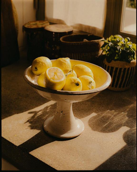 A white ceramic bowl filled with lemons sits on a counter, illuminated by sunlight creating strong shadows.