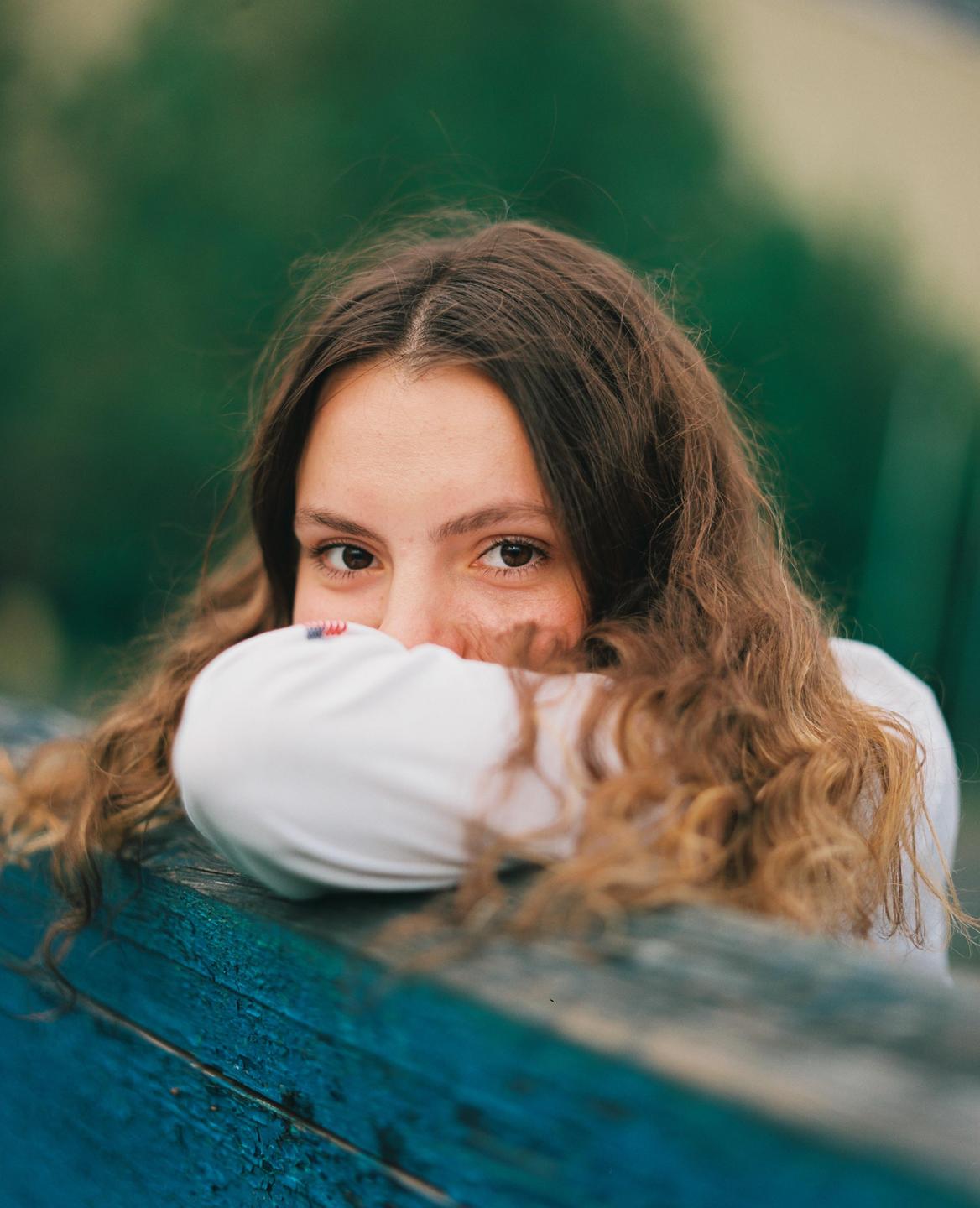 A woman with long, curly hair rests her arms on a blue wooden surface, looking directly at the viewer.