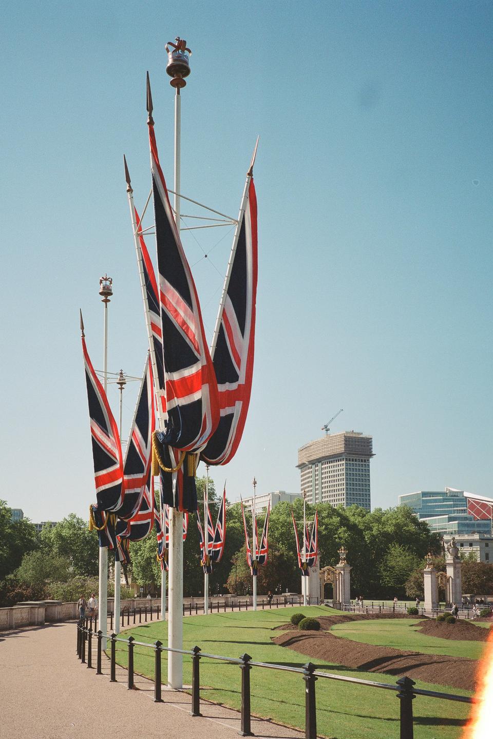 Union Jack flags hang from poles along a paved path next to a green lawn with a building in the background.