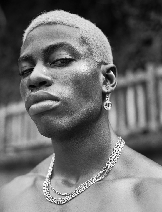 A man with short, light-colored hair, an earring, and multiple necklaces looks directly at the viewer.
