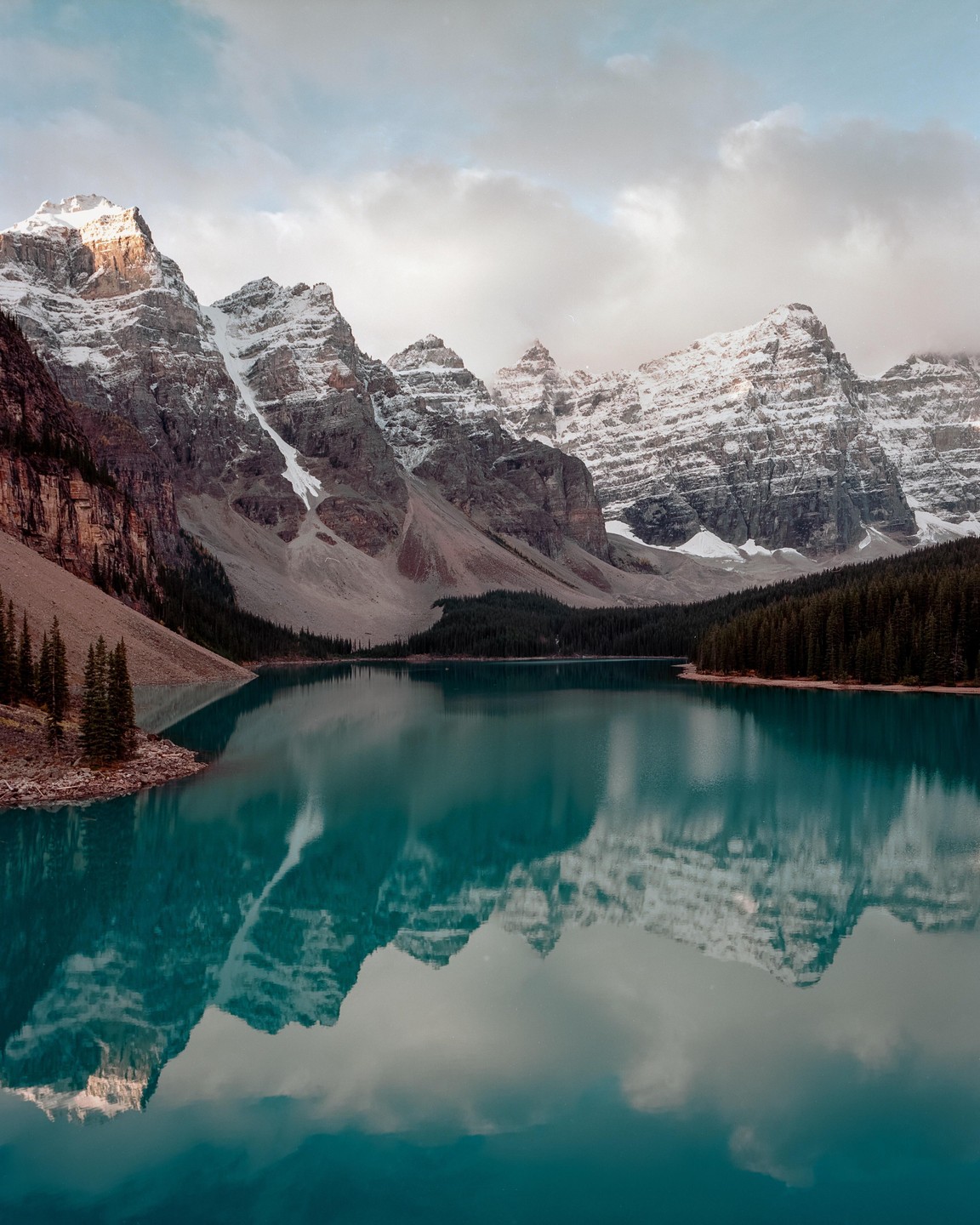 Snow-capped mountains are reflected in a turquoise lake surrounded by a pine forest under a cloudy sky.
