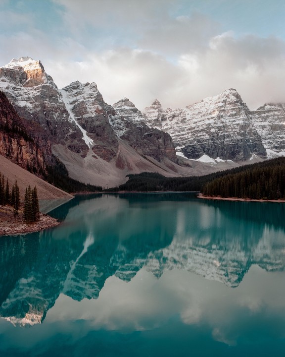 Snow-capped mountains are reflected in a turquoise lake surrounded by a pine forest under a cloudy sky.