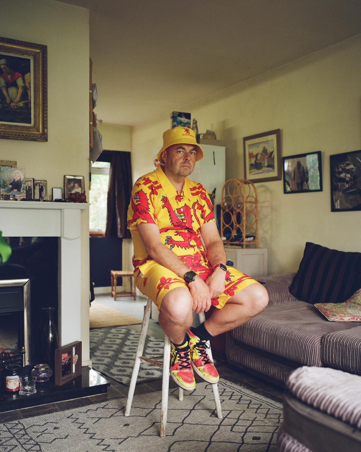 A man wearing a yellow and red outfit with a lion rampant pattern sits on a stool in a living room.