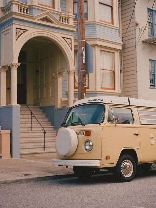 A beige camper van is parked on a street in front of a house with an arched entrance.