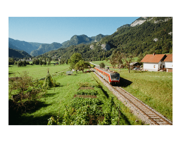 A red train travels along a track through a green valley with mountains and houses in the background.