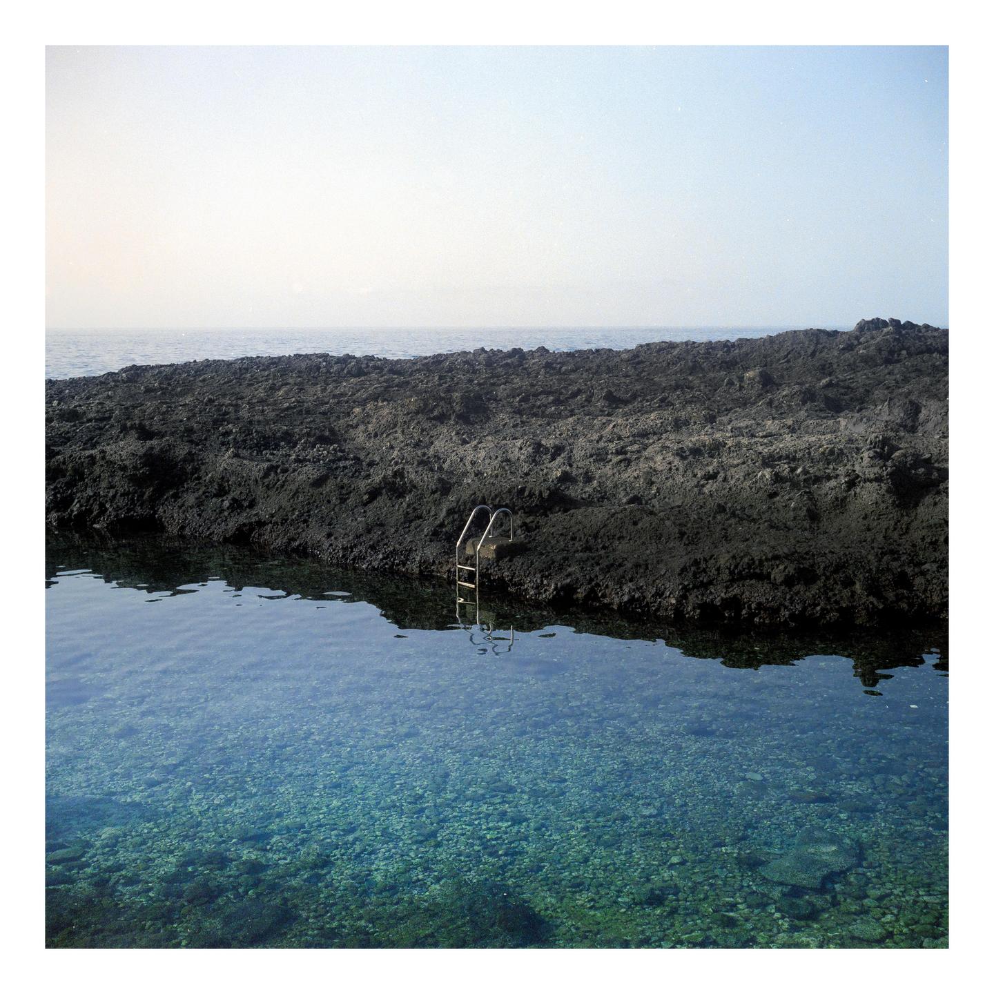A metal ladder extends into a clear blue natural pool surrounded by dark volcanic rock with the ocean in the distance.