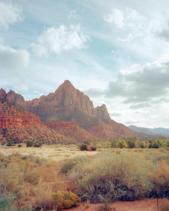 A vast landscape features a prominent red rock mountain under a cloudy sky, with dry brush and green foliage in the foreground.
