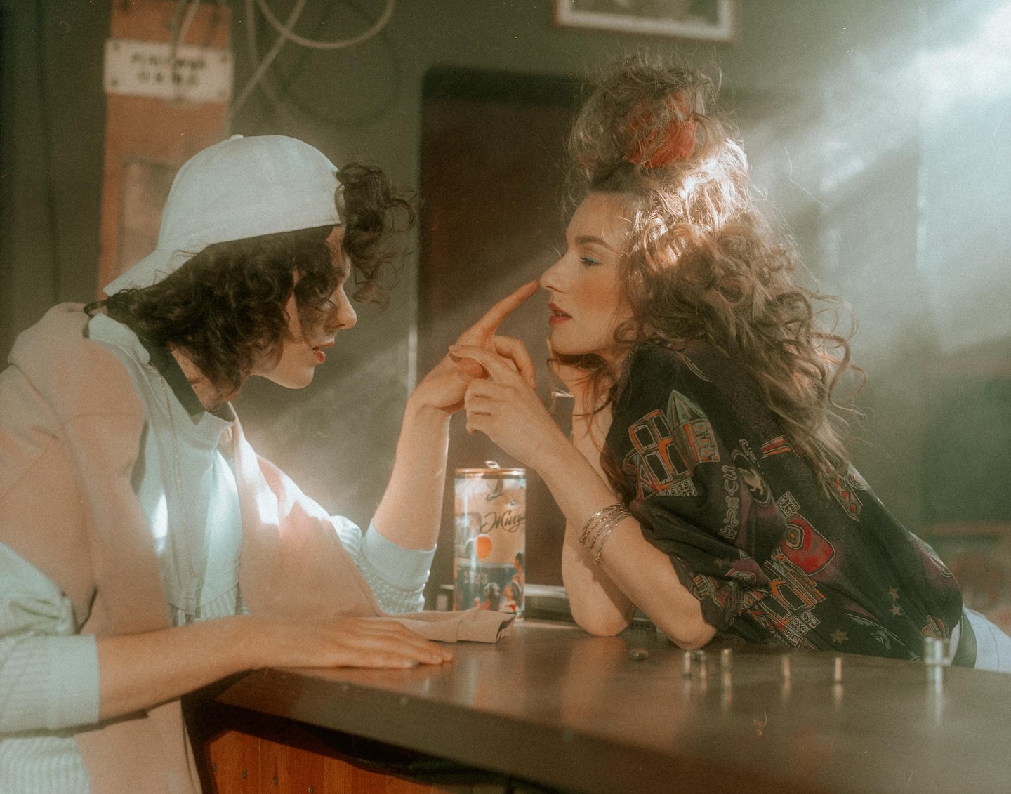 A woman in a baseball cap and a woman with curly hair are sitting at a bar, with the woman in the cap touching the other woman's nose.