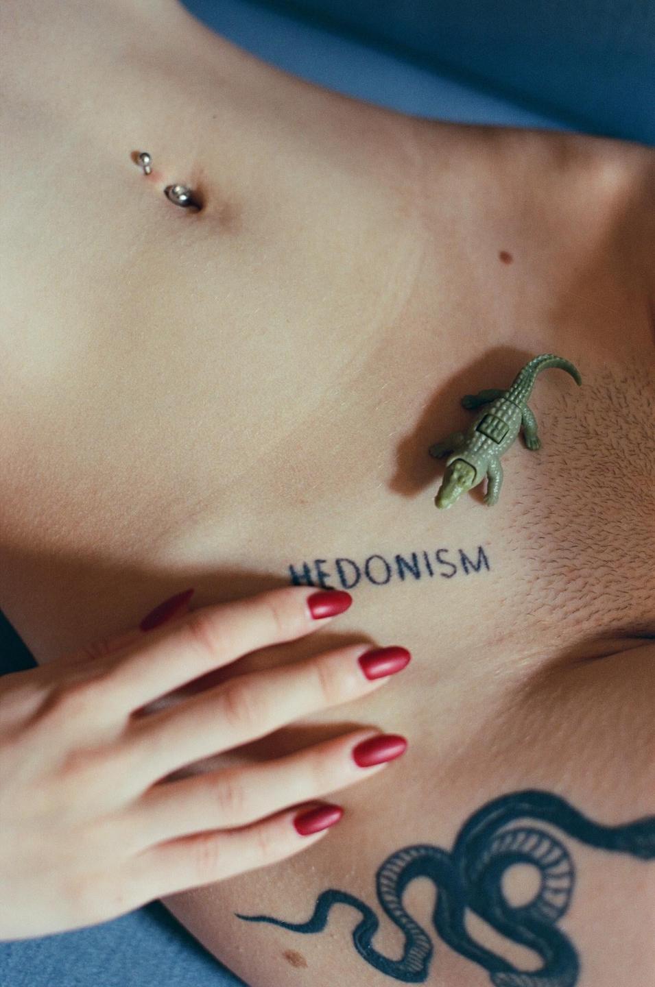 A person's torso is visible with a belly button piercing, a tattoo that says "HEDONISM", a snake tattoo, a small toy alligator, and a hand with red fingernails.
