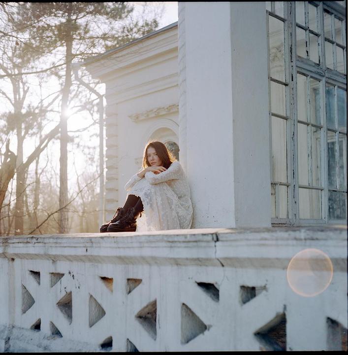 A woman in a white dress and boots sits on a ledge next to a white building with trees in the background.