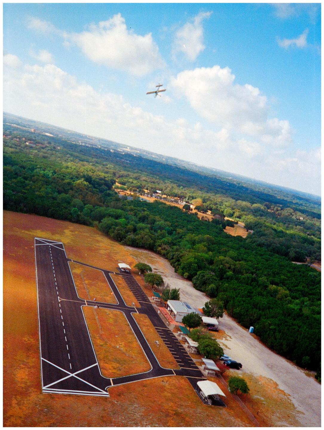 An aerial view shows a small airplane flying above an airfield with a runway and surrounding trees.