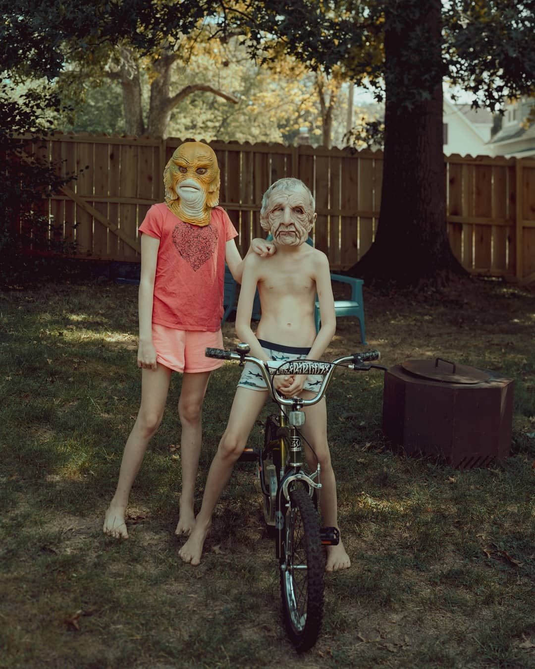 Two children wearing monster masks stand barefoot in a grassy backyard, one with a bicycle.