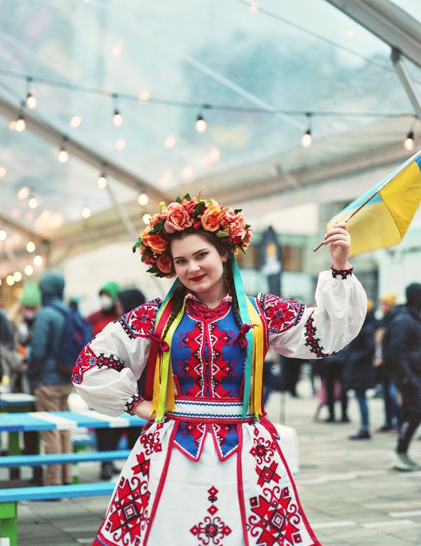 A woman in traditional Ukrainian dress and a floral crown holds a small Ukrainian flag under a tent with string lights.