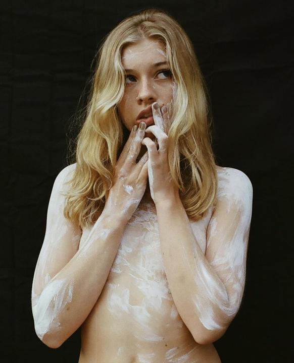 A woman with long blonde hair and white paint on her body looks to the side against a dark background.