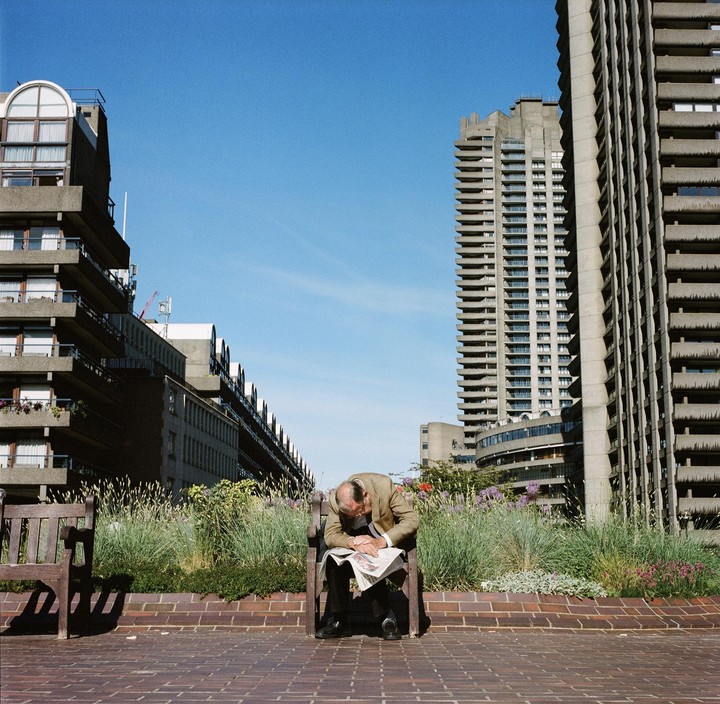 A man sleeps on a bench with a newspaper on his lap, surrounded by tall concrete buildings and green plants.