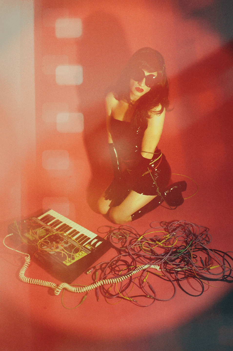 A woman in a shiny black outfit and sunglasses kneels on a red surface next to a synthesizer and a pile of cables.