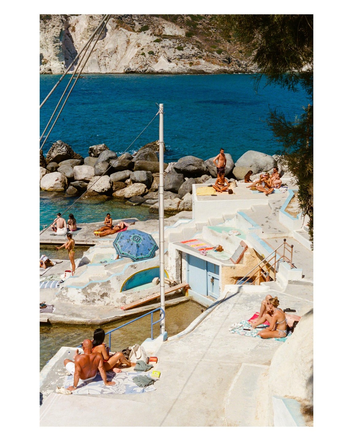 People relax on concrete platforms and rocks next to a body of blue water with a rocky cliff in the background.
