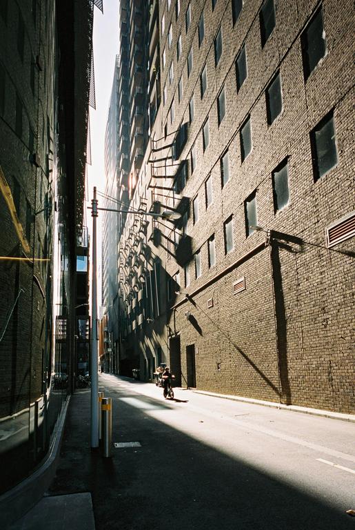 A person rides a bicycle down a narrow alley between tall brick buildings, with sunlight creating strong shadows on the right wall.
