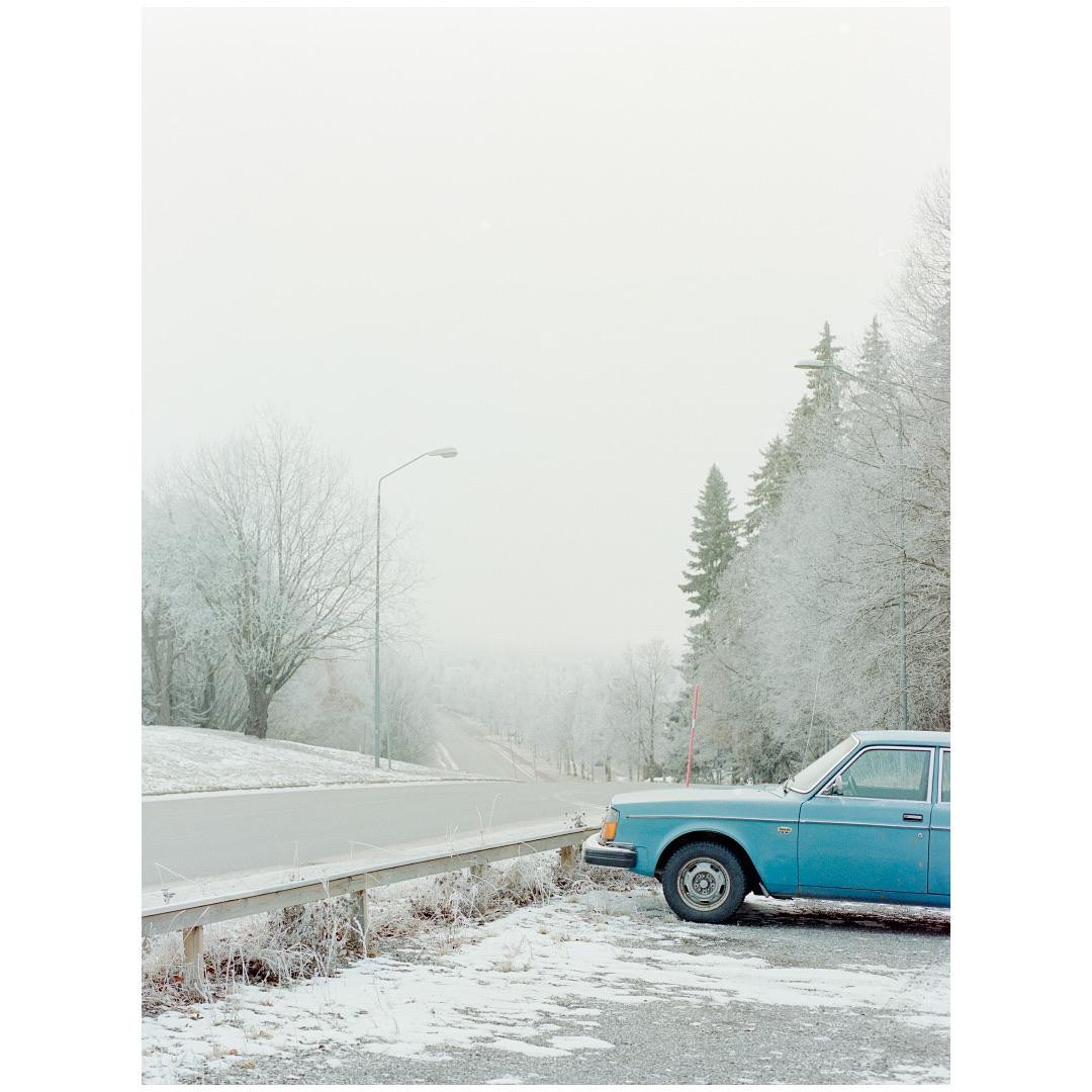 A blue car is parked on the side of a snowy road next to a guardrail, with frost-covered trees and a street light in the background.