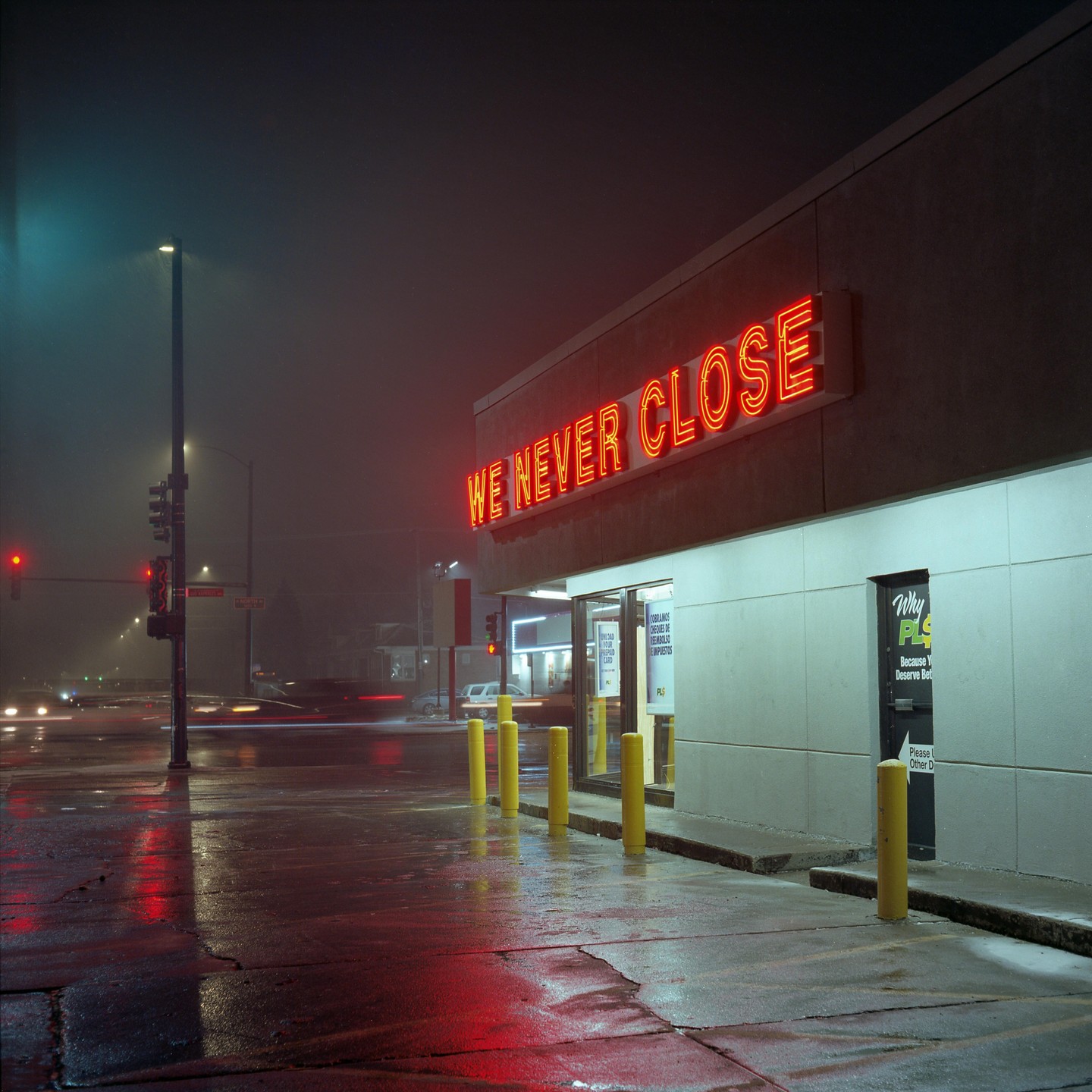 A building with a neon sign that reads "WE NEVER CLOSE" stands next to a wet street at night.