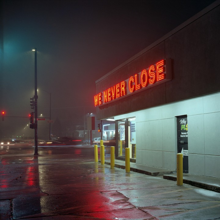 A building with a neon sign that reads "WE NEVER CLOSE" stands next to a wet street at night.