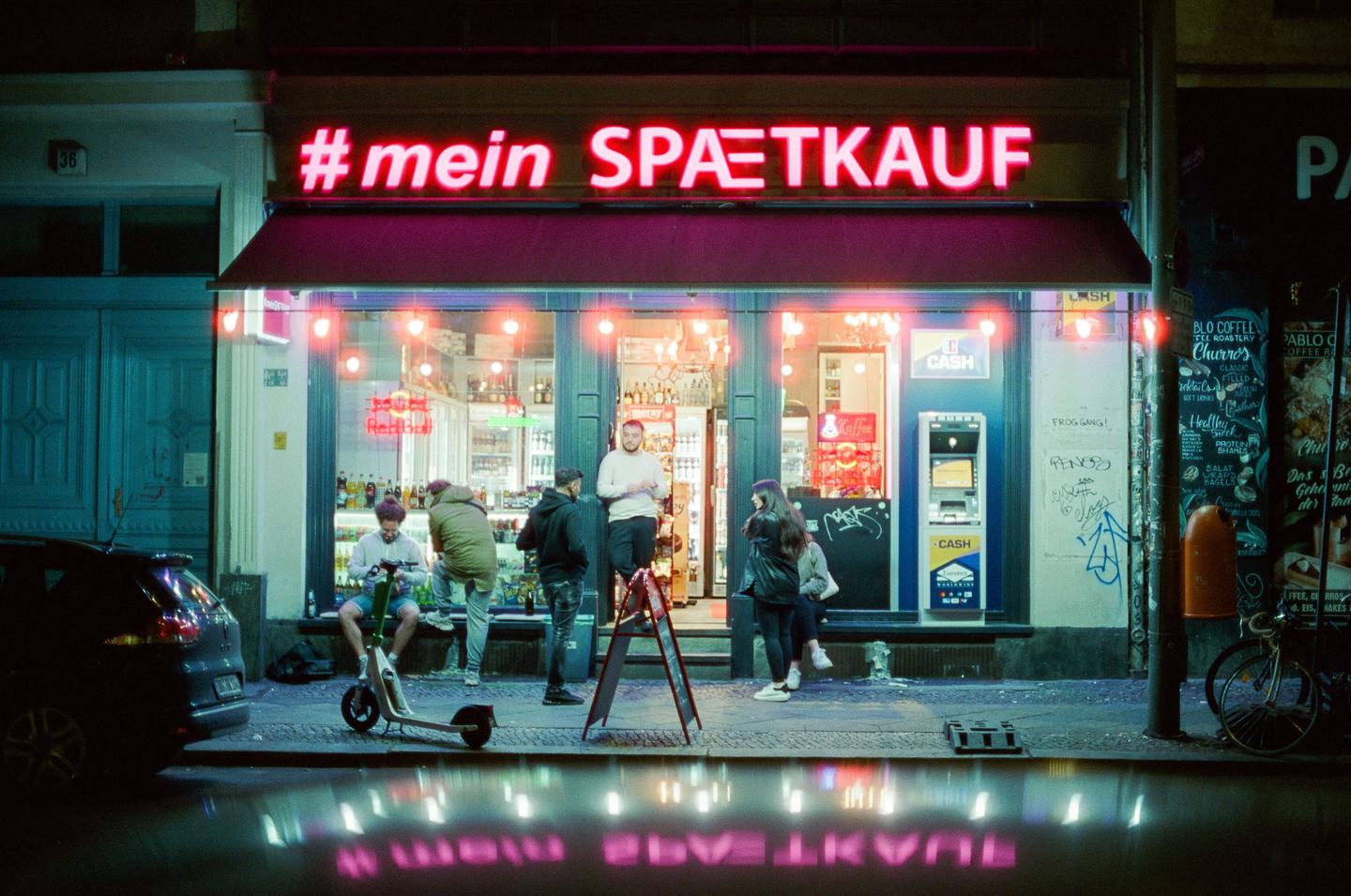 A group of people stand outside a convenience store with a neon sign that reads "#mein SPAETKAUF" at night.