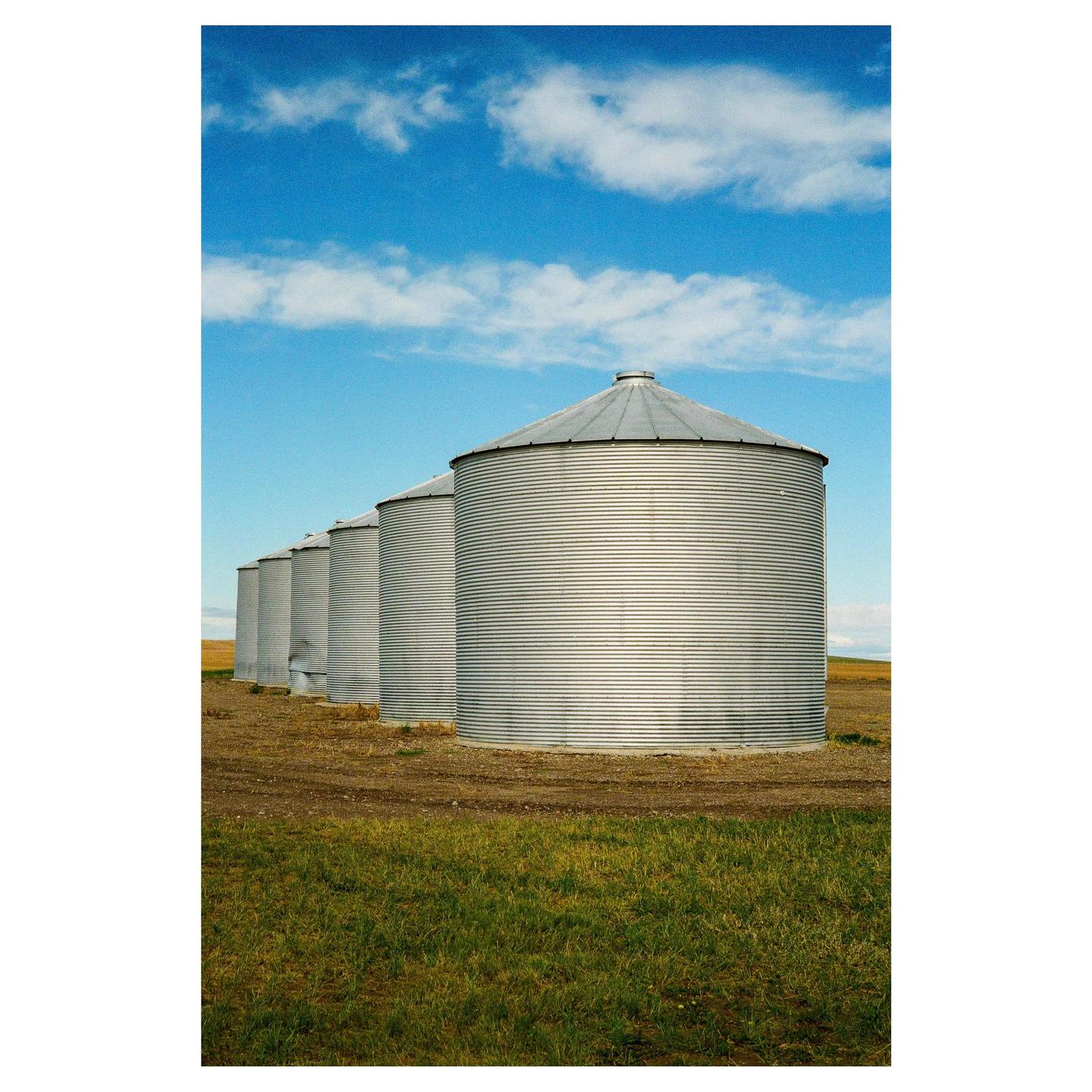 A row of grain silos stands in a field under a blue sky with white clouds.