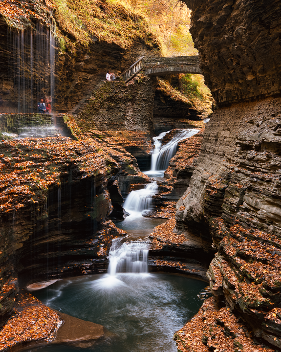 A series of waterfalls cascade through a rocky gorge covered in autumn foliage, with a stone bridge and people visible in the upper left.