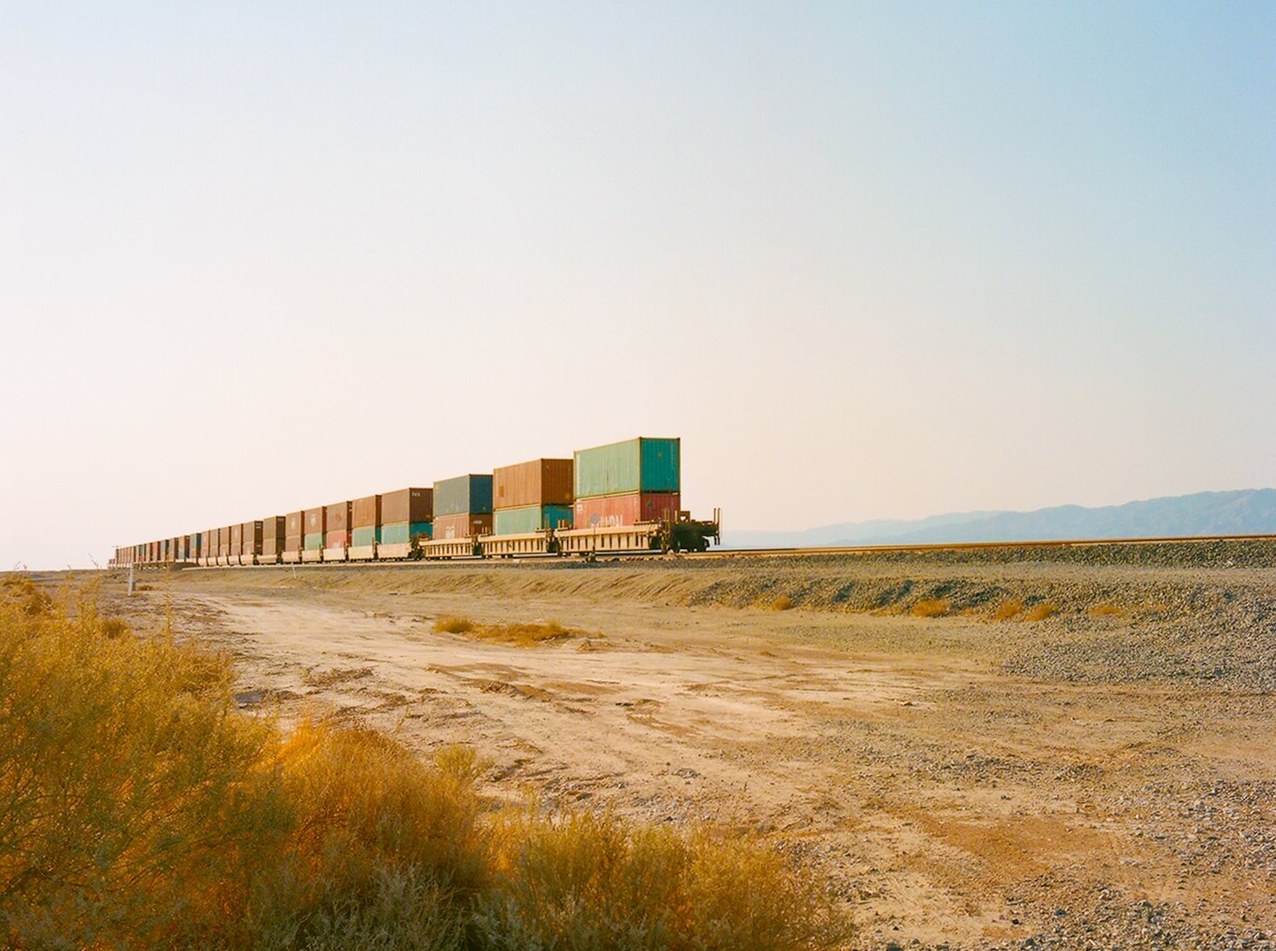 A long freight train with colorful containers travels through a desert landscape under a clear sky.