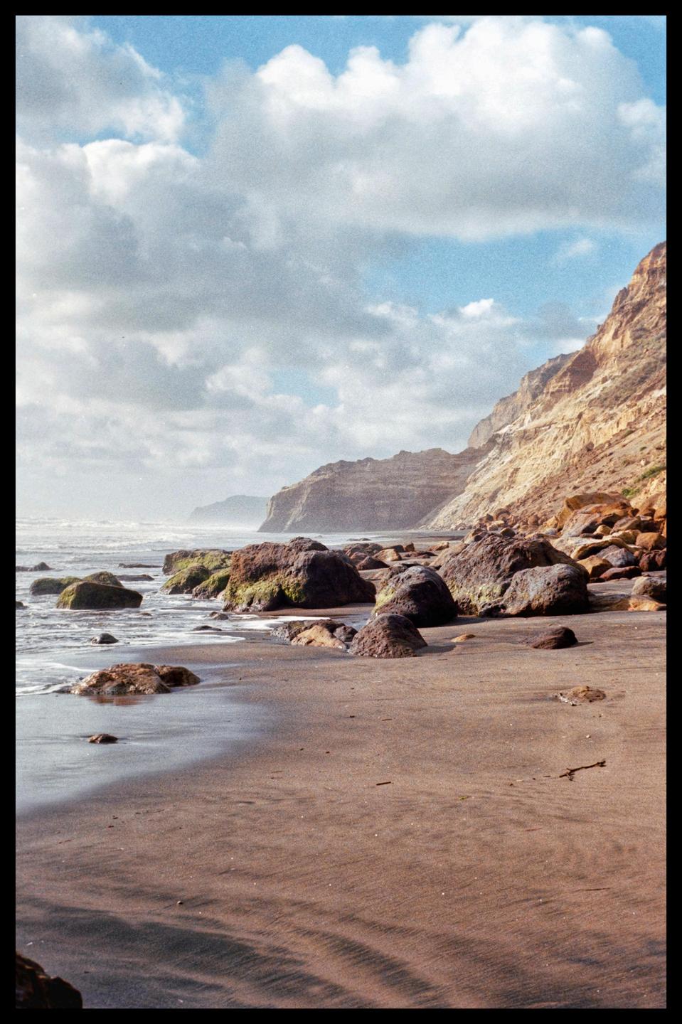 Waves crash on a rocky beach with cliffs in the background under a cloudy sky.
