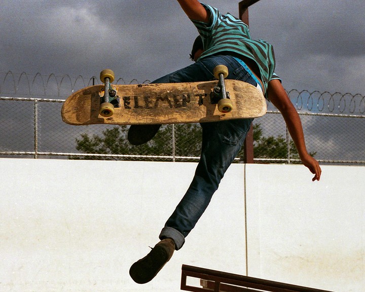 A person in a striped shirt and jeans is airborne on a skateboard with a fence and trees in the background.