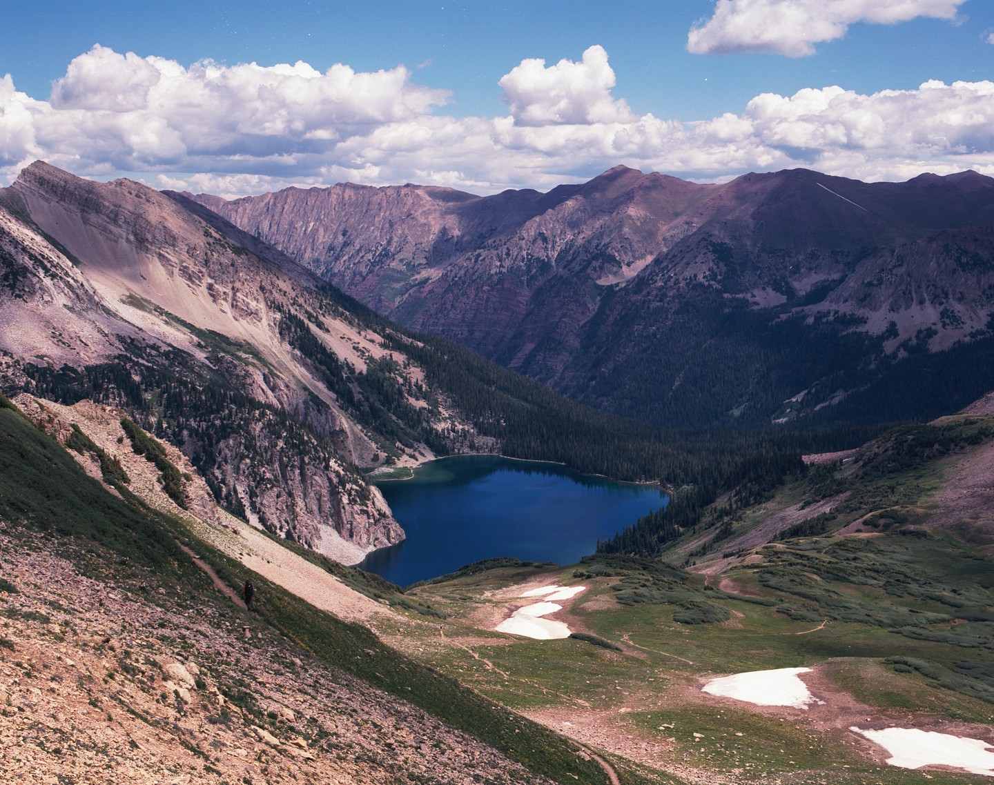 A deep blue lake is nestled in a valley surrounded by mountains with patches of snow and green foliage under a cloudy sky.