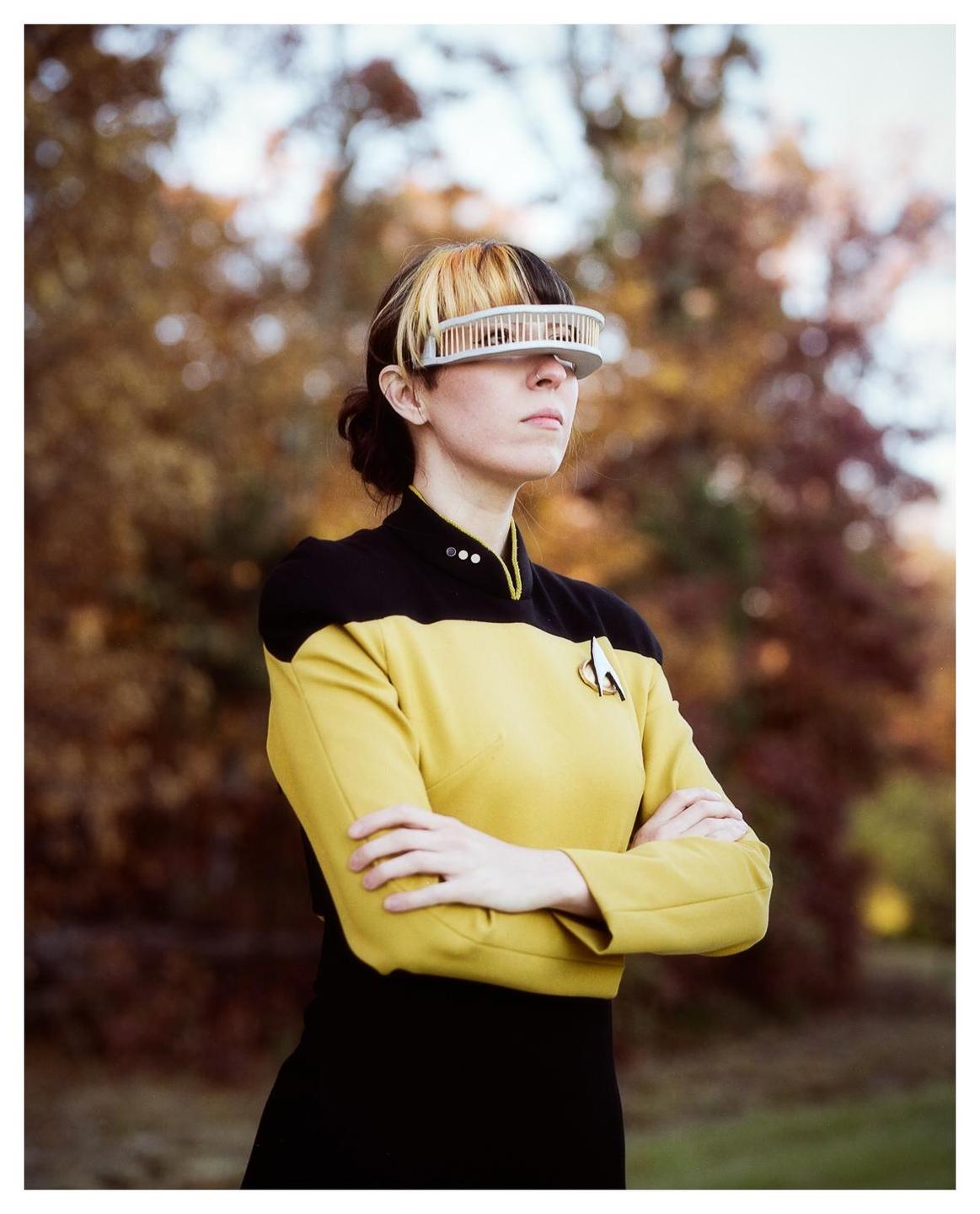 A woman in a yellow and black uniform with a futuristic visor stands with her arms crossed outdoors.