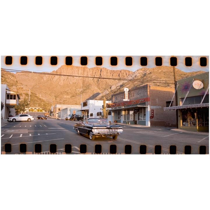 A classic convertible drives down a street lined with buildings and a mountain in the background.