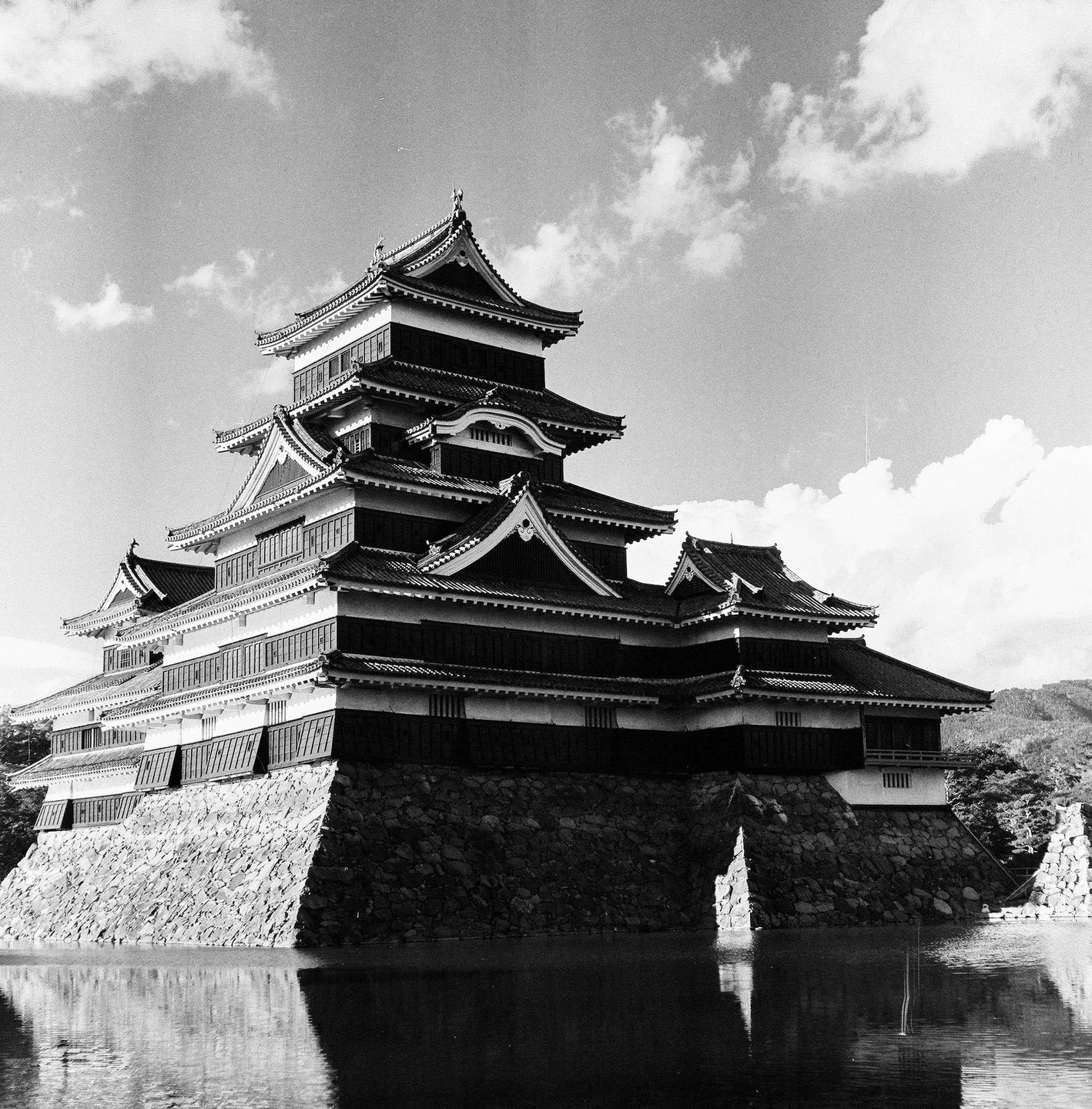 A large, multi-story Japanese castle with a dark facade and white trim stands on a stone foundation surrounded by water, with mountains in the background.