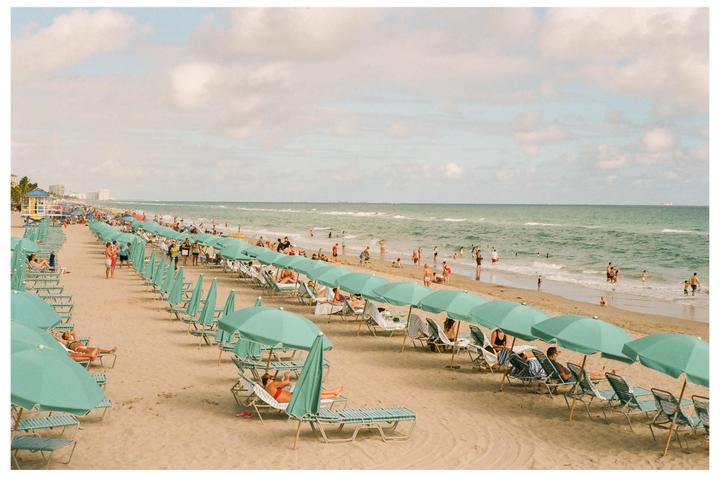A long line of teal beach umbrellas and lounge chairs stretches along a sandy beach with people swimming and relaxing.