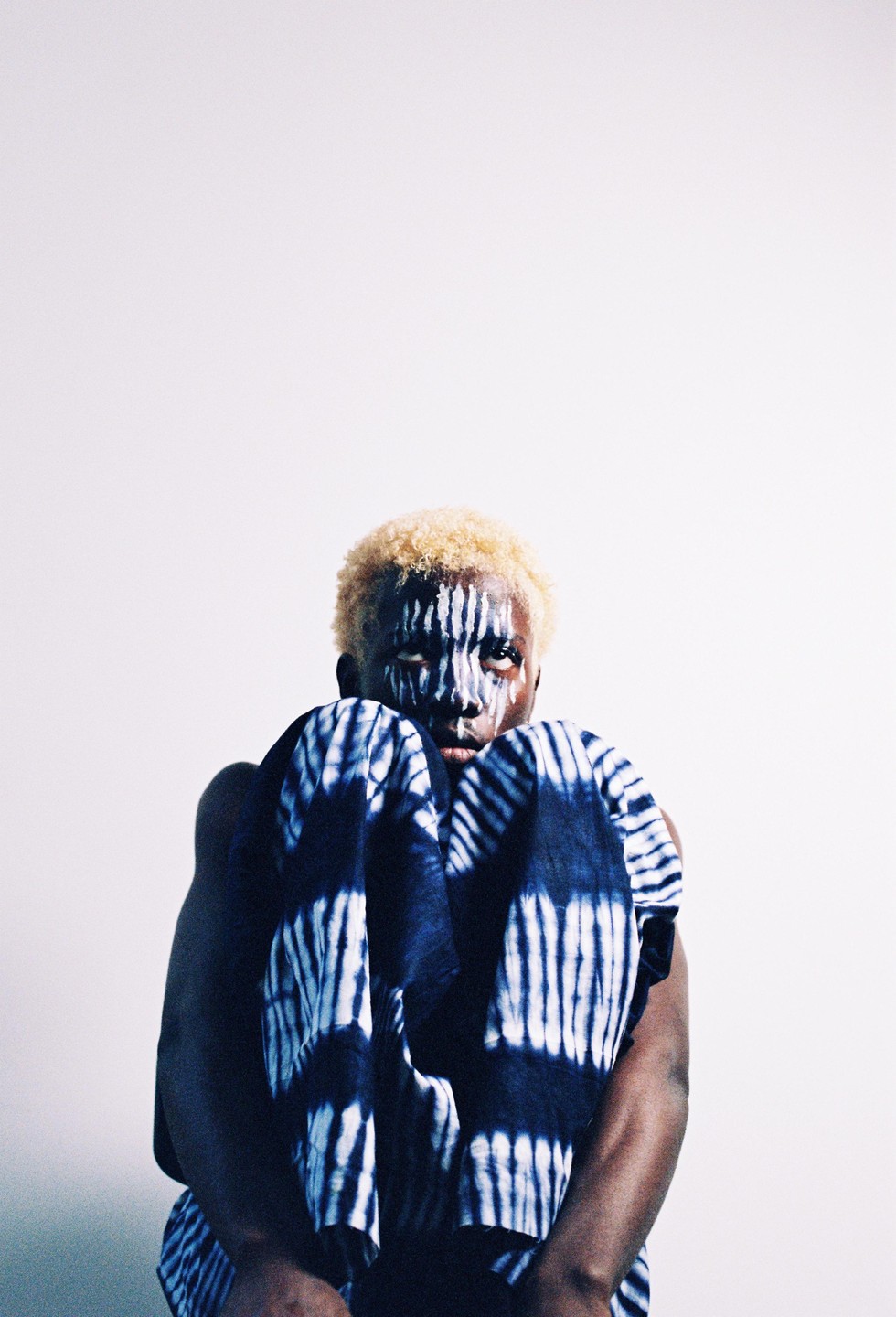 A person with bleached hair and white face paint looks directly forward while holding a blue and white patterned fabric.