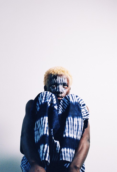 A person with bleached hair and white face paint looks directly forward while holding a blue and white patterned fabric.