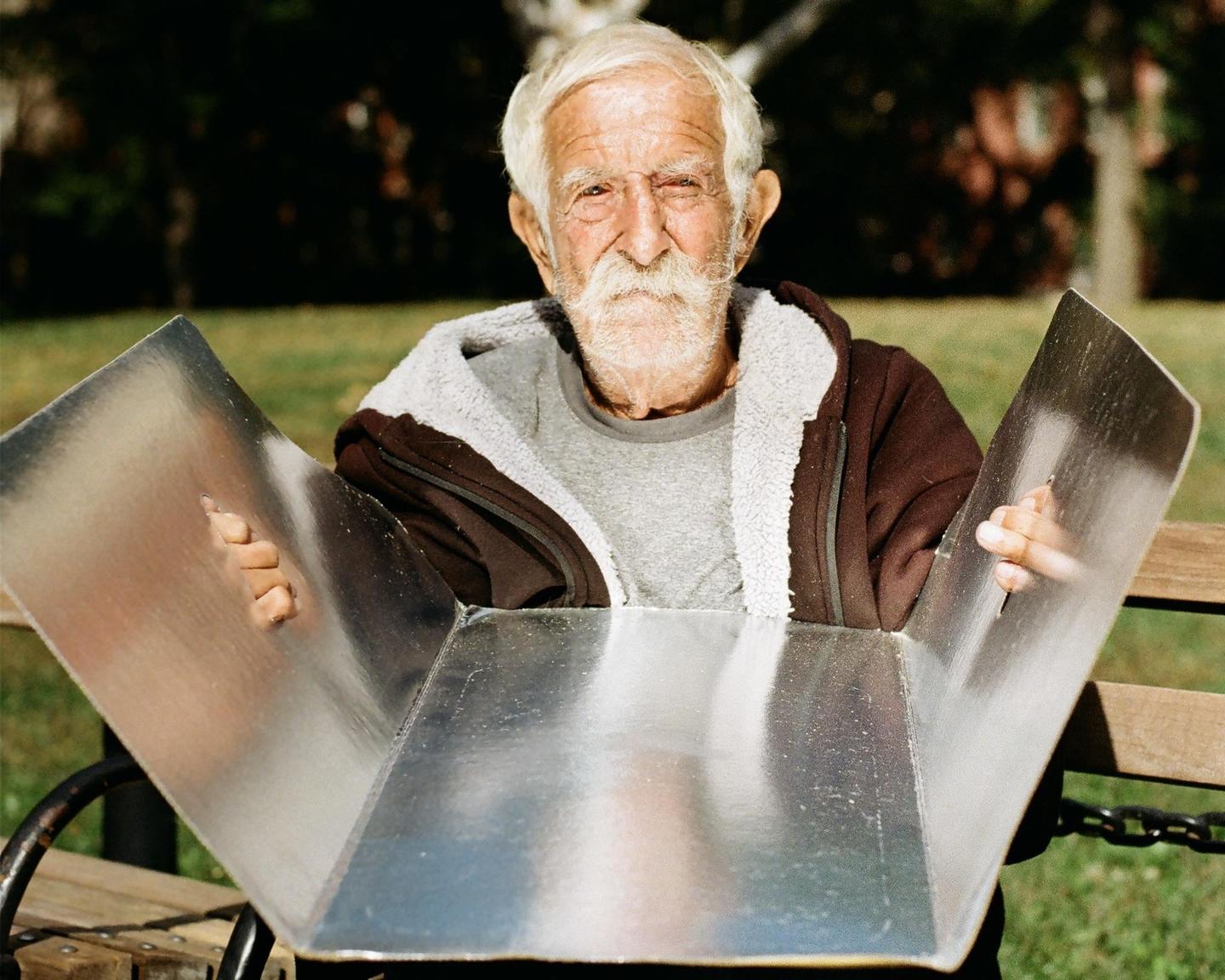 An elderly man with a white beard holds a large metal reflector in a park.