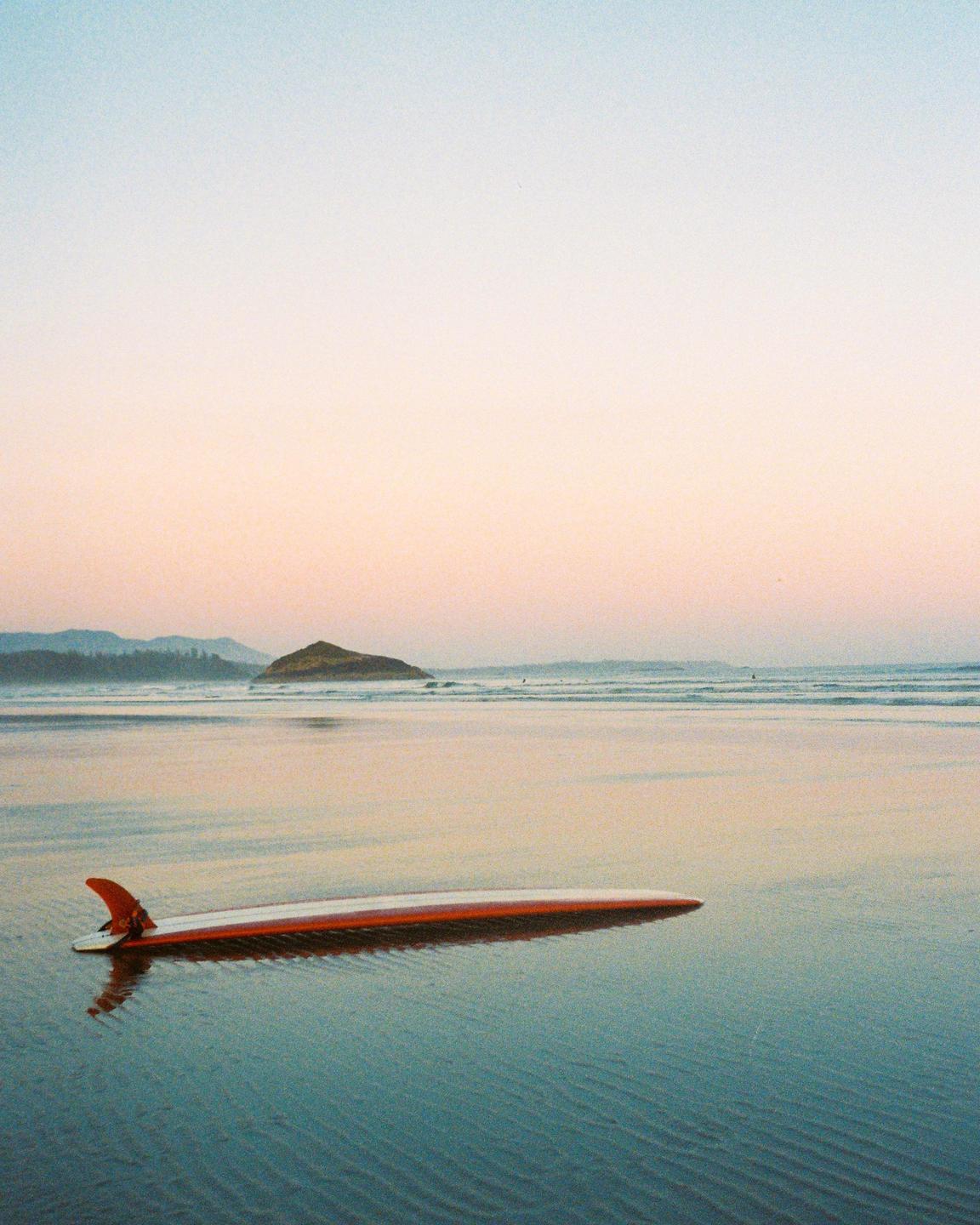 A surfboard rests on the wet sand of a beach with an island in the distance under a pastel sky.