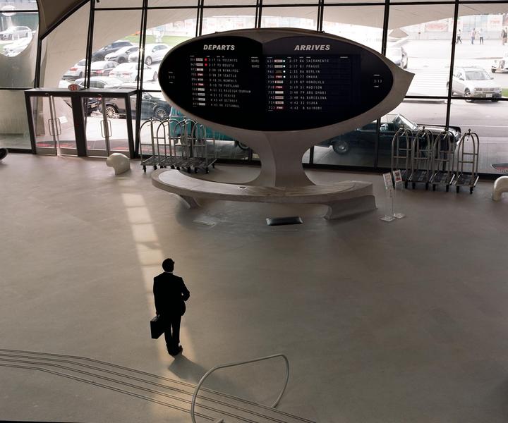 A man in a suit with a briefcase stands on a light-colored floor in front of a large departure and arrival board.