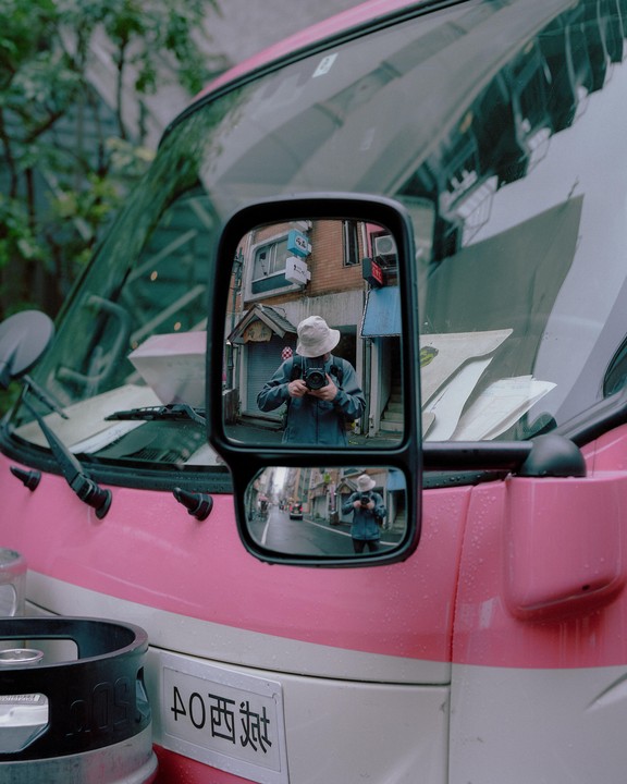A man wearing a bucket hat takes a self-portrait in the side mirror of a pink and white vehicle on a street in Tokyo.
