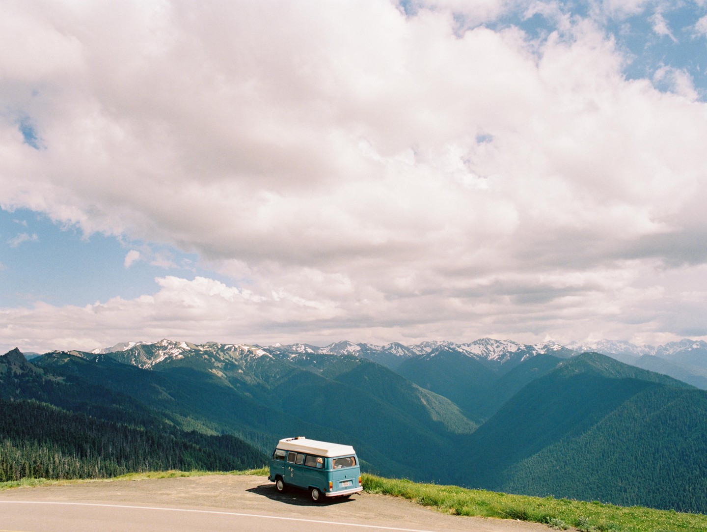 A blue van is parked on the side of a road overlooking a vast mountain range with snow-capped peaks under a cloudy sky.