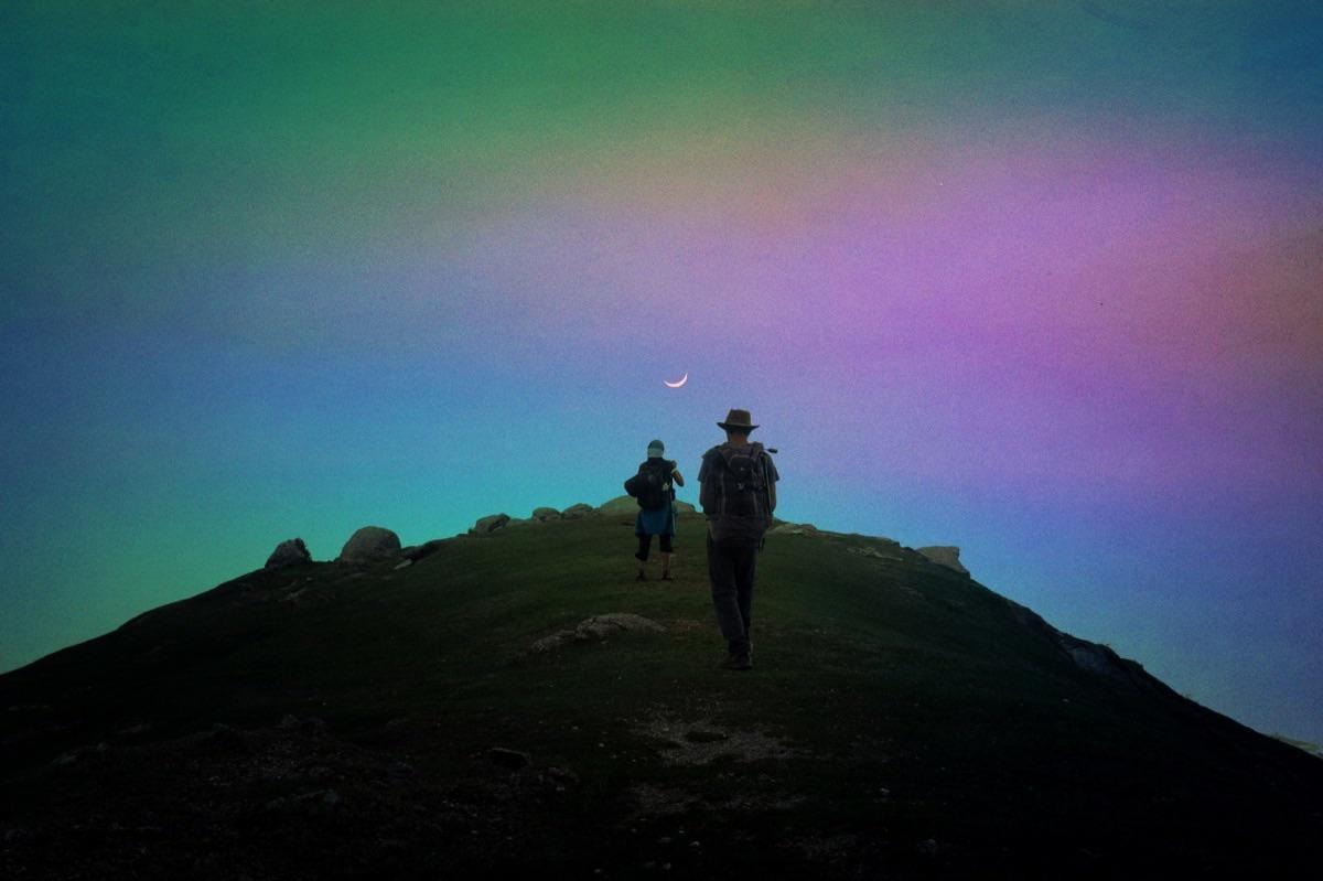 Two people with backpacks walk up a grassy hill under a colorful sky with a crescent moon.