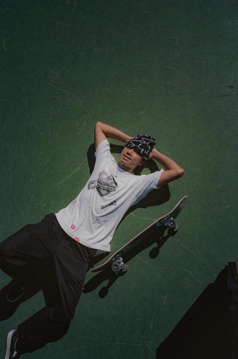A young man wearing a beanie and t-shirt lies on a green surface with a skateboard next to him.