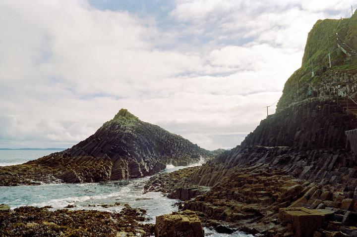 A body of water flows between two large, dark, rocky formations under a cloudy sky.