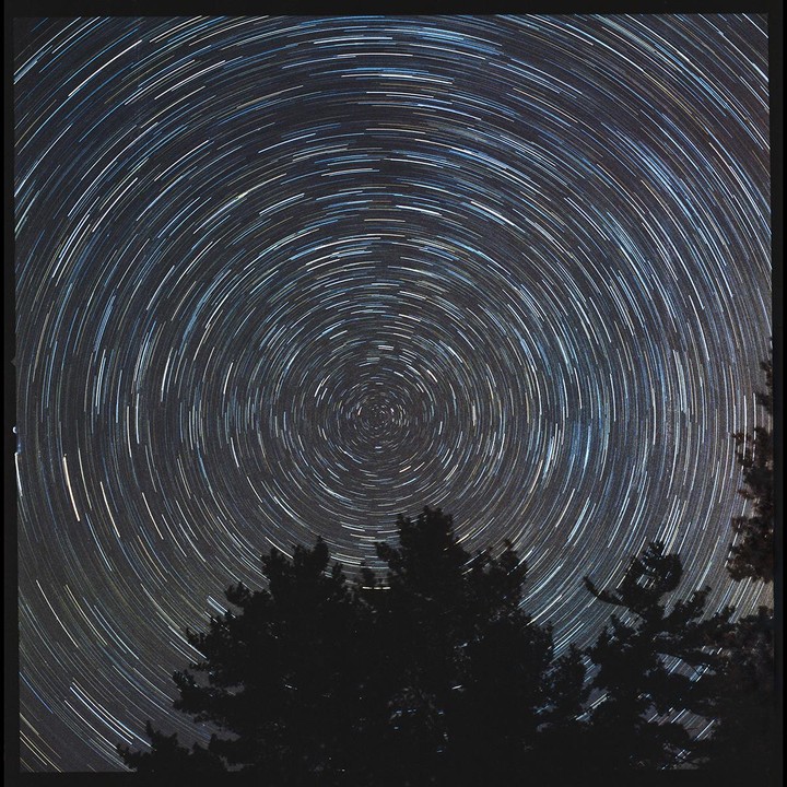 The night sky shows star trails circling the North Star above a dark tree line.