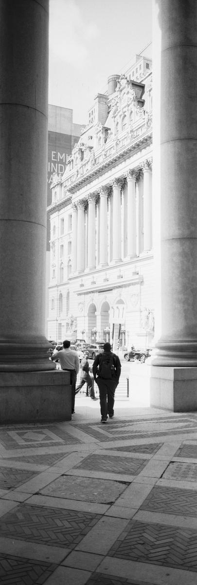 Three men walk on a patterned sidewalk between two large columns, with a classical building and street visible in the background.